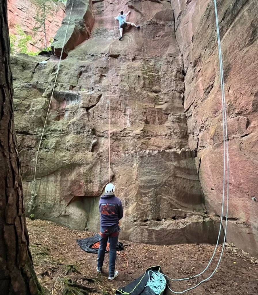 Ein Kletterer an der Sandsteinwand ist im Vorstieg unterwegs. Der Sichernde steht unten und sieht mit Helm auf dem Kopf nach oben. 